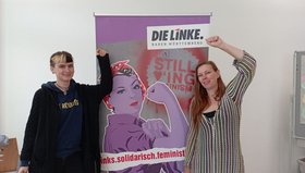 Anna Damoune und Isabell Fuhrmann stehen links und recht von einem Roll-Up mit der Aufschrift "link.solidarisch.feministisch"